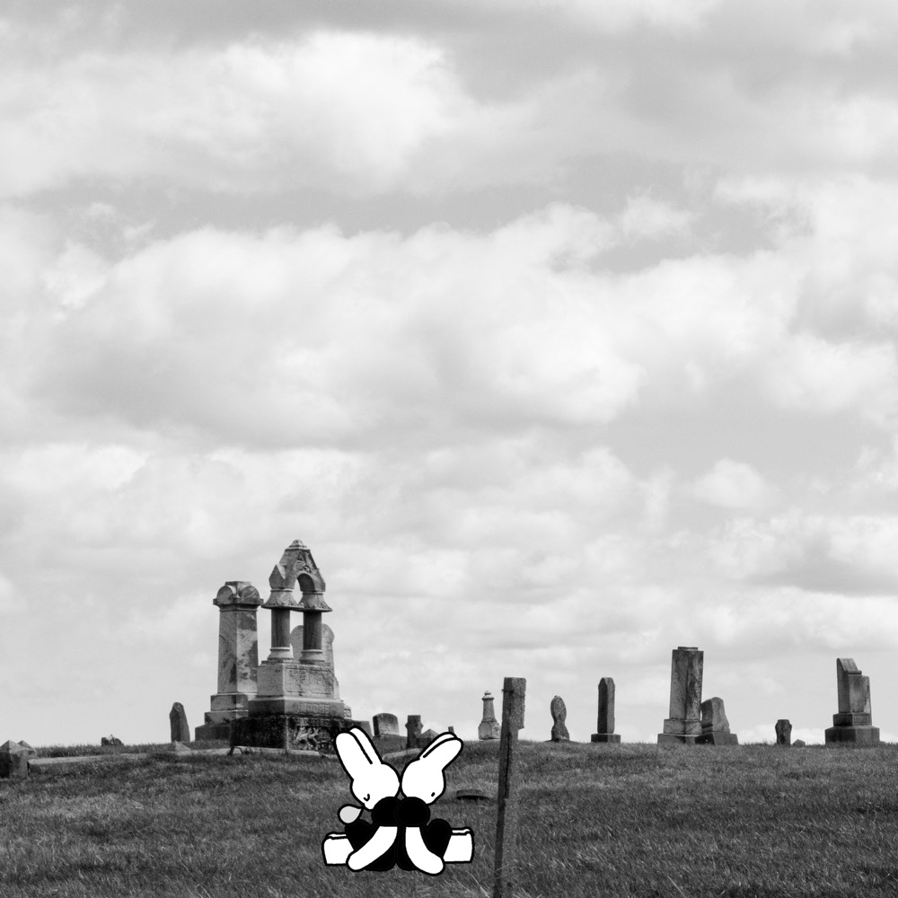 Black and white image, two animated white rabbits sit with their backs facing one another in the foreground. The background hosts a cemetary. Image by Soft and Dumb.