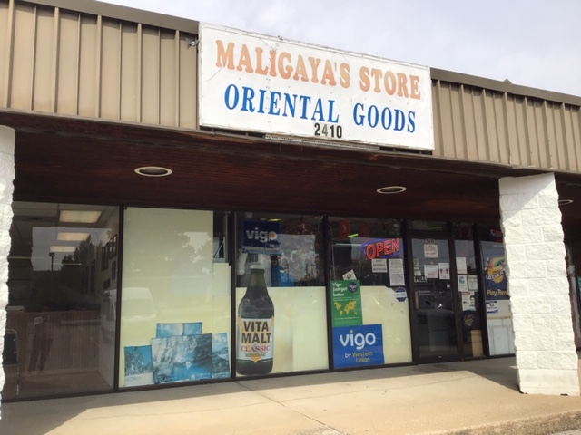 A view of the simple sign which reads â€œMaligayaâ€™s Store Oriental Goodsâ€ on a sunny day. The  store is located in a shopping center with a brown metal awning supported by white rough brick columns. The sign sits on the brown awning; beneath it in the storefront windows the red and white â€œOpenâ€ sign is lit. The windows also contain a few advertisements for some of their products and services. Photo by Rachael McMillan.