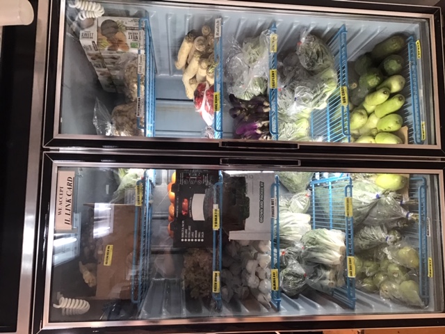 A double-door glass cooler contains neatly stacked and labeled fresh produce. There are green onions, garlic, tomatoes, radishes, long beans, chinese cabbage, and bottle gourds, among many other varieties of fresh vegetables. A small sign taped to the top left door reads â€œWE ACCEPT IL LINK CARD.â€ Photo by Rachael McMillan.