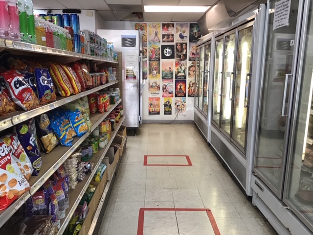 A view down an aisle: one side contains shelving with packaged chips, jarred/canned fruits, and prepared drinks. At the end of the aisle is a double-door cooler with specialty items; they arenâ€™t visible from the outside. The other side of the aisle contains see-through coolers/freezers filled with meat, seafood, and other perishable items. The view terminates in a view of a wall with slightly larger than notebook-sized posters of movies available for rent. Photo by Rachael McMillan.