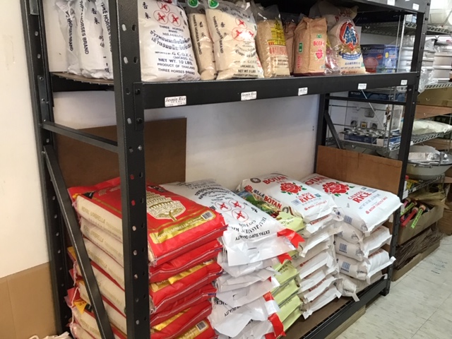 Dark metal industrial shelving holds large sacks of rice in different varieties on the bottom shelf, with smaller bags on the top shelf. Photo by Rachael McMillan.