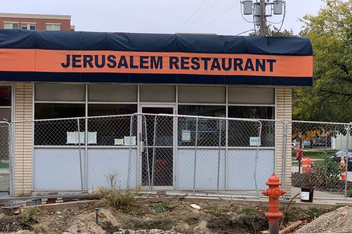 Photo of Jerusalem Restaurant from across the street. An orange and blue awning reads â€œJerusalem Restaurantâ€ in blue writing. The building bricks are cream colored and there are two windows on either side of the center door. There is a chain link fence in front of the restaurant because of local construction. Photo by Megan Friend.