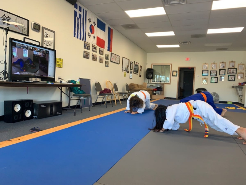 3 kids holding a plank position at Kokushi judo in front of a TV during a Zoom judo clinic with 3x Olympic bronze medalist Amarilis Savon. Two students are wearing white uniforms, one blue is wearing a blue uniform, all with orange belts. The flooring is mainly grey and blue, with a yellow stripe. There are four flags hanging on the wall the students are facing. Photo by Grace Talusan.