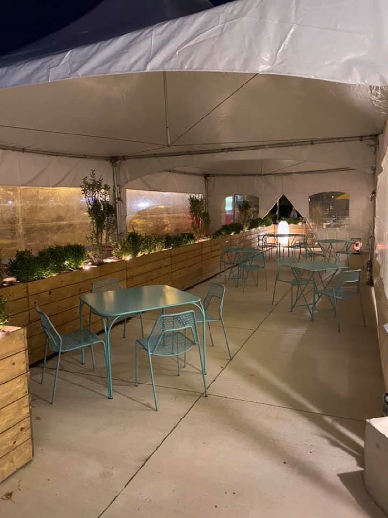 Several blue-green metal tables are spread out across a patio. There is greenery along one side, and a large white tent covering. Photo from Everyday Kitchen Facebook page.
