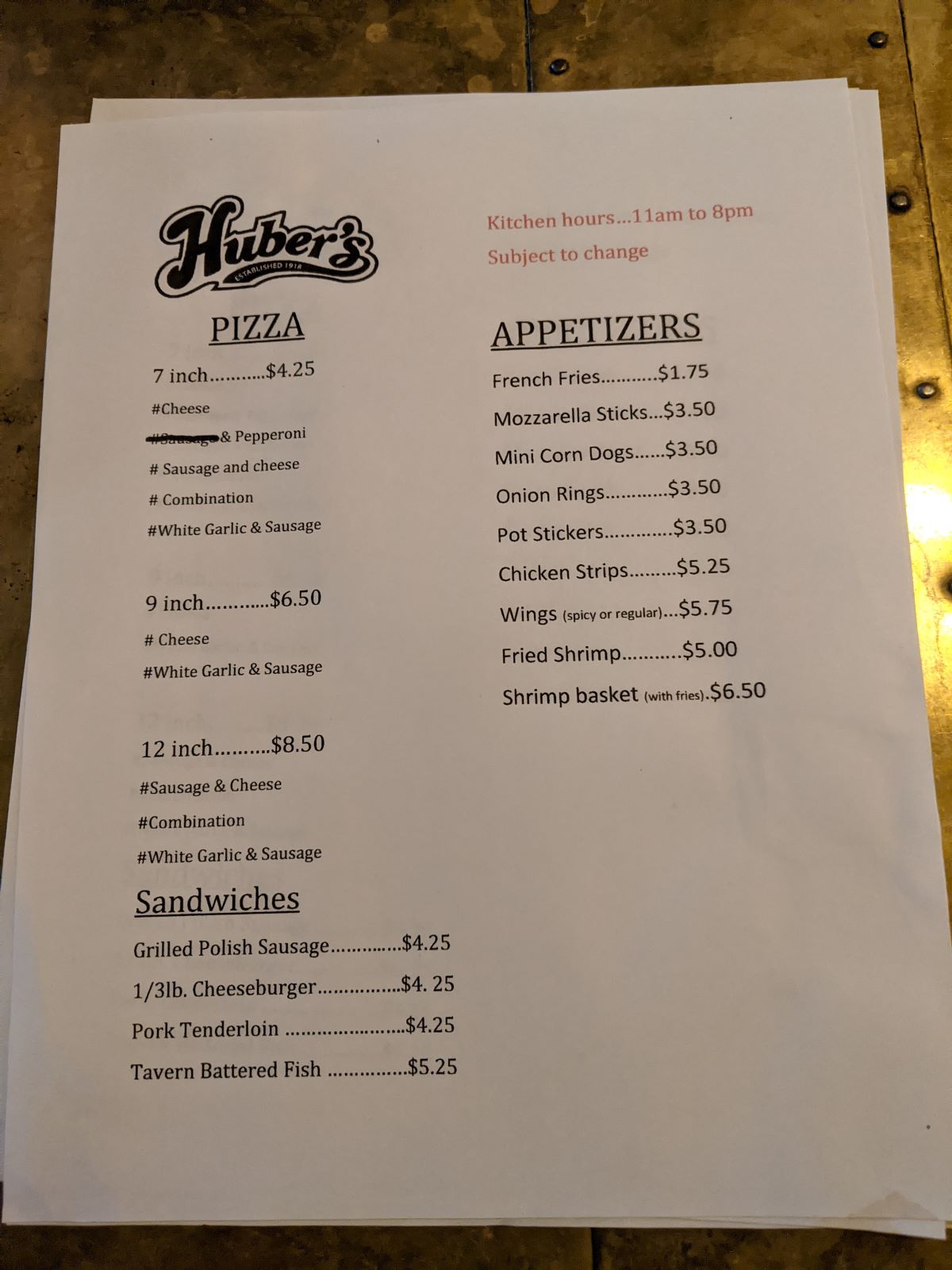 A paper menu of food offerings and their prices, including pizza, sandwiches, and appetizers.  Kitchen hours are listed in red at the top right hand corner of the menu. Photo by Tias Paul.