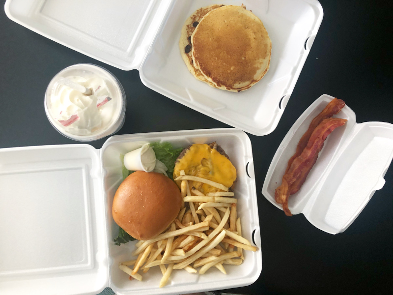 An overhead shot of a delivery order from Merry Ann's Diner featuring a mikshakes, a burger and fries in an open styrofoam container, another container holding panckes, and a skinny styrofoam container holding two slices of bacon. Photo by Alyssa Buckley.