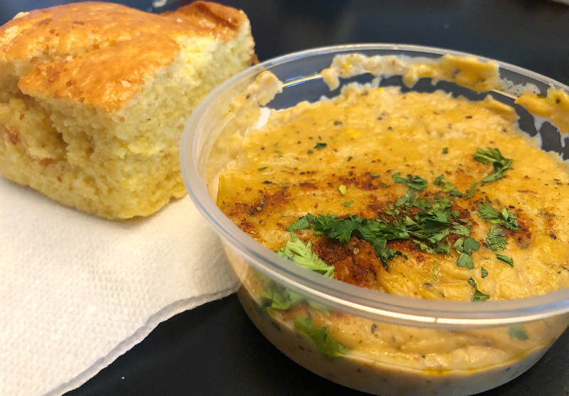 In a small takeout container, there is an orange colored bisque from Neil St. Blues in Champaign. There is a large square of cornbread on a white paper napkin beside it. Photo by Alyssa Buckley.
