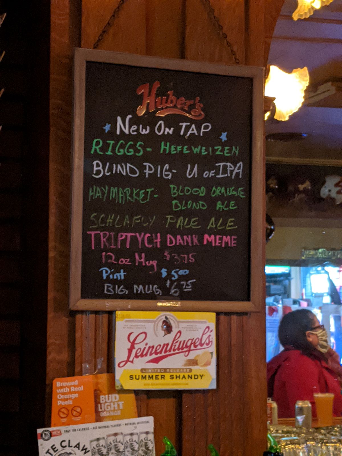 A wood-framed black chalkboard sign listing the local draft beers that are available on tap.  A yellow and white â€œLeinenkugelâ€™sâ€ sign is directly below the chalkboard.  To the right, we can see a mirror reflection of a masked patron sitting at the bar with a plastic cup of beer. Photo by Tias Paul.