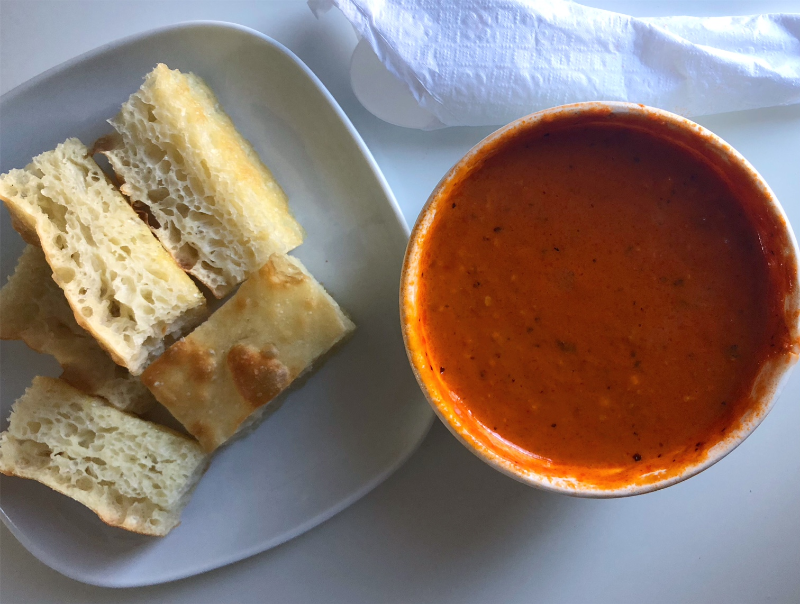 An overhead photo of red pepper soup from Pekara Bakery on Springfield in Champaign. The soup is in a to go cup, and beside it, there is a white square plate with five pieces of bread. Photo by Alyssa Buckley.