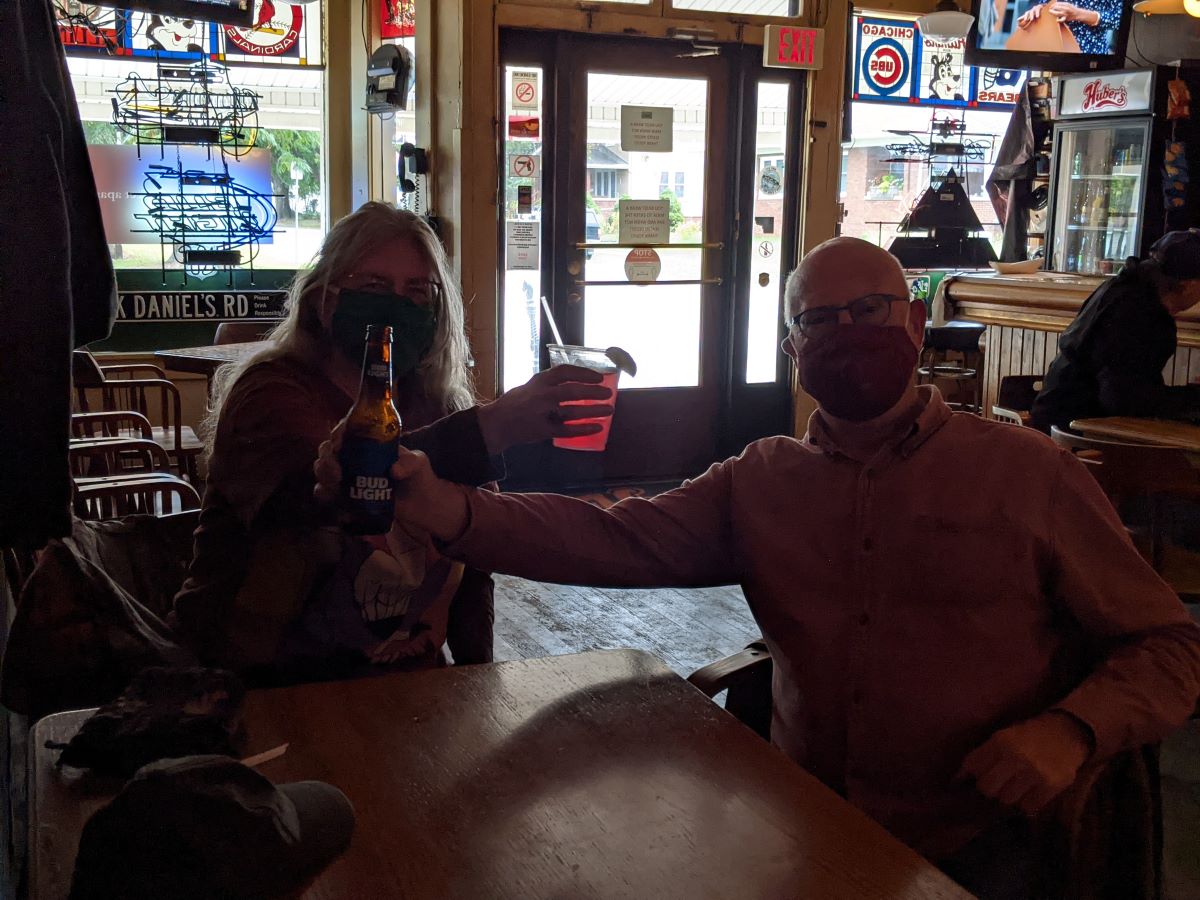 A picture of two Huberâ€™s patrons wearing masks, each holding up a drink.  They are sitting at a wooden table close to the bar area.  Behind them, we see the main entrance to the store, looking out onto Church St. Photo by Tias Paul.
