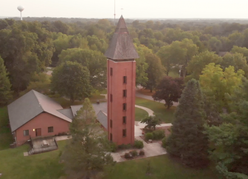 a very tall bell tower is seen from above amongst a forest; a water tower is in the background