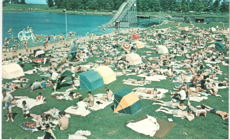 hundreds of people from the 1960s congregate on a grass beach; a massive waterslide is in the background
