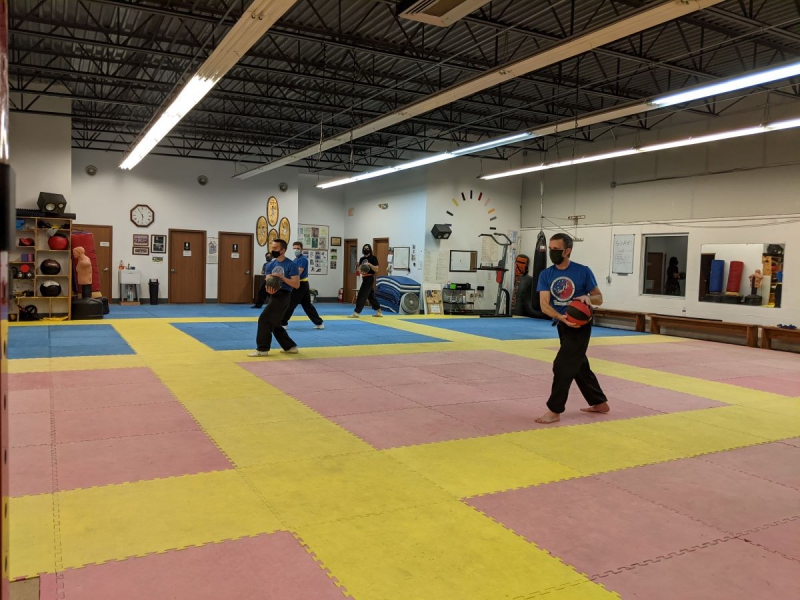 Instructor David Ward (front) and Songâ€™s students holding medicine balls during the practice of a qigong exercise. Most of the students are wearing blue T-shirts and black pants. The floor is made of pink, yellow, and blue exercise mats. The room is lit by rows of fluorescent lighting visible by the ceiling. Photo by Tias Paul.