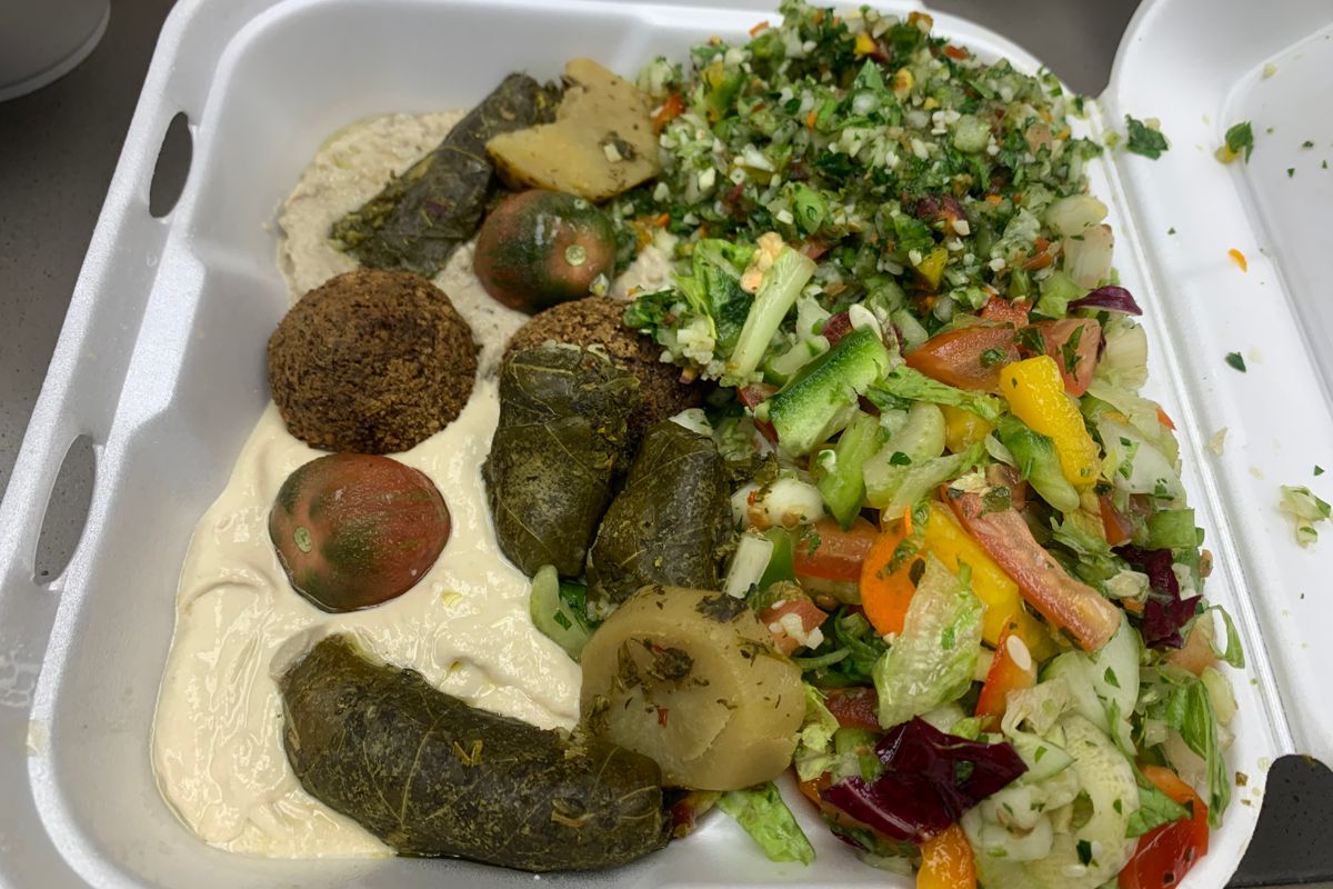 A photo of a Styrofoam takeaway container with falafel balls, stuffed grape leaves, cherry tomatoes and a few sliced potatoes sitting on top of hommoss and baba ghanouj dips on the left. Next to that are a tabbouleh salad with chopped herbs, tomato and cucumber and fatouch salad with sliced lettuce, bell pepper, tomato and herbs. Photo by Megan Friend.