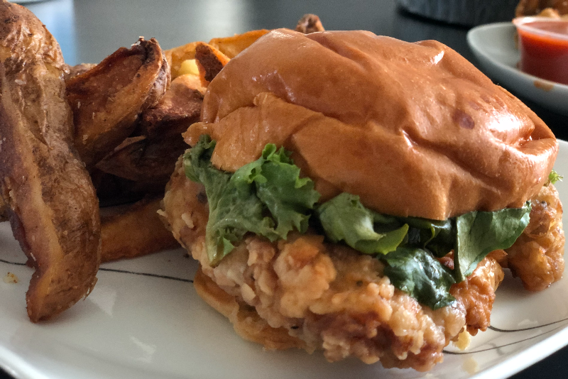 A fried chicken sandwich from Watson's sits on a white plate next to potato wedges. Photo by Alyssa Buckley.