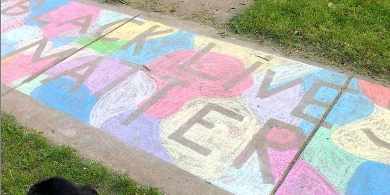A multi colored chalk drawing with the words Black Lives Matter in negative relief on a sidewalk. There is grass on either side of the sidewalk, and a black dog's head at the bottom of the image. Photo by Jessica Hammie. 