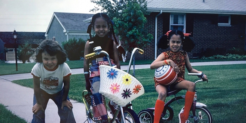 Three young girls look at the camera, two on the right are sitting atop bicycles.