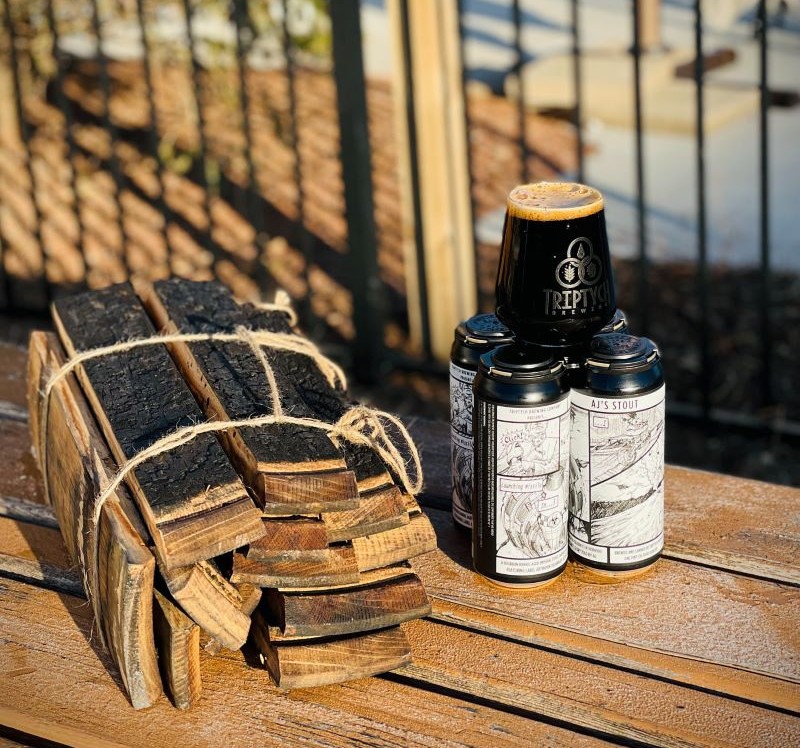 A four-pack of black and white beer cans are sitting on a picnic table. There is a glass of dark beer sitting on top of them. To the left is a bundle of wooden barrel half staves. Photo from Triptych Brewing Facebook page.