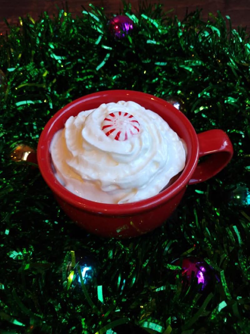 A red mug has a thick swirl of whipped cream and a round peppermint candy in the center. It sits on a backdrop of green shiny tinsel. Photo from Aroma Cafe Facebook page. 