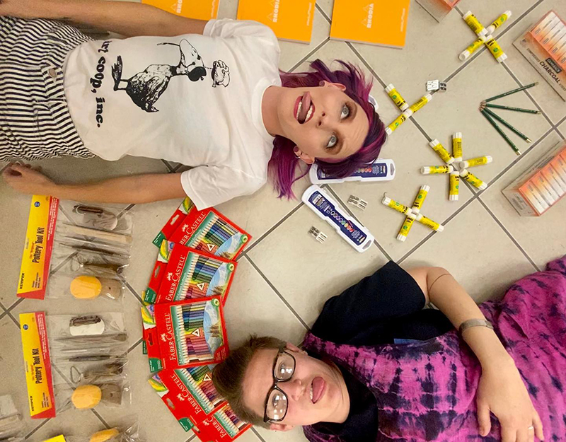 Two employees of art coop lay on the ground with the camera view directly above them. They are surrounded by art supplies. Photo from Art Coop's Facebook page.