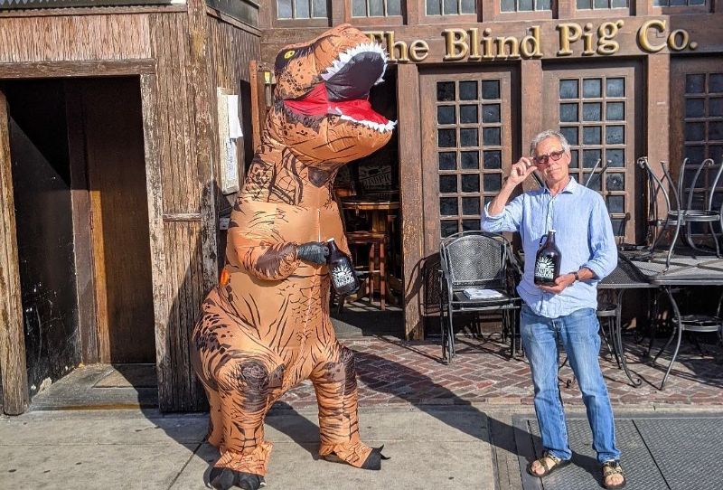 In front of Blind Pig, there is a giant, inflatable T-rex costumed person holding a growler next to a white man holding a growler. The T-rexâ€™s mouth is open as if roaring, and the man is touching his glasses with one hand and is wearing Teevas. Behind them, there is one chair set up and an outdoor table with chairs stacked upside down on top of them. 