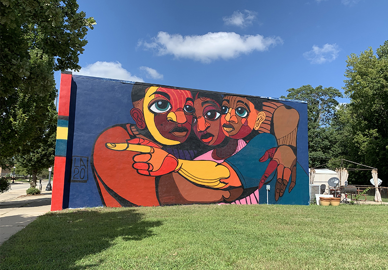 A mural with three bodies hugging one another, colored in blue, green, yellow, and various other colors. Photo by Debra Domal.