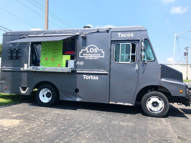 Los Hidalguense taco truck, a black food truck, is parked in the parking lot of Hunter Haven in Champaign. The exterior of the truck has white lettering which reads the name of the truck in addition to â€œTacosâ€ and â€œTortas.â€ The lime green menu is visible on the side of the truck, and the sky behind it is a bright, cloudless blue. 