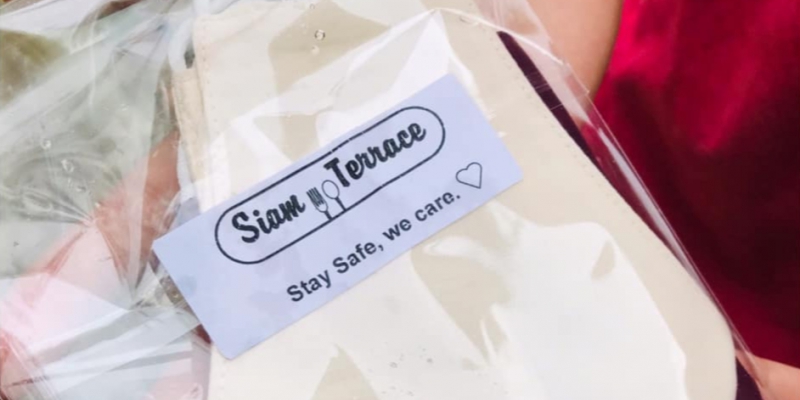 The photo is a close up of Siam Terrace in Urbanaâ€™s free white face masks. It is packaged in a clear, plastic wrapper with a white sticker reading, â€œSiam Terrace. Stay safe, we care.â€ plus a small heart. Photo from Siam Terraceâ€™s Facebook page.