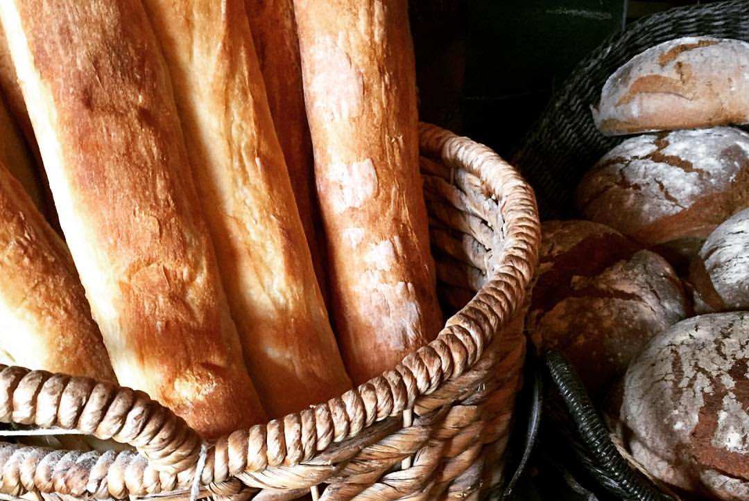 Many fresh baguettes are in a tall wicker basket beside another basket of sourdough rolls. Photo from The Bread Company's Facebook page.