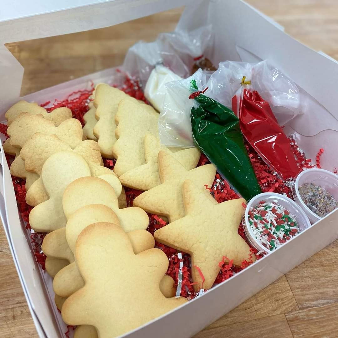 In a white paper box, there are 13 unfrosted sugar cookies in holiday shapes next to four bags of icing and containers of sprinkles. This is a cookie decorating set from Central Illinois Bakehouse. Photo from Central Illinois Bakehouse Facebook page.