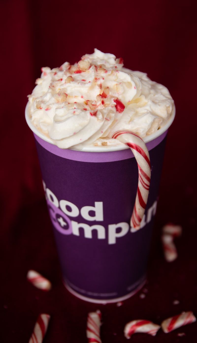 A deep purple paper cup is topped with whipped cream and crushed peppermints. There is a candy cane hanging off the edge of the cup. The cup says 