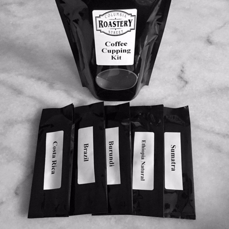 Five small coffee bags are laid in a row with white labels of the coffee's origin. Behind the small bags there is a larger black bag reading 