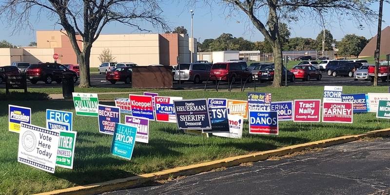 Political yard signs are clustered in a grassy parking lot divider. Beyond the signs is a parking lot full of cars. Photo by Julie McClure. 