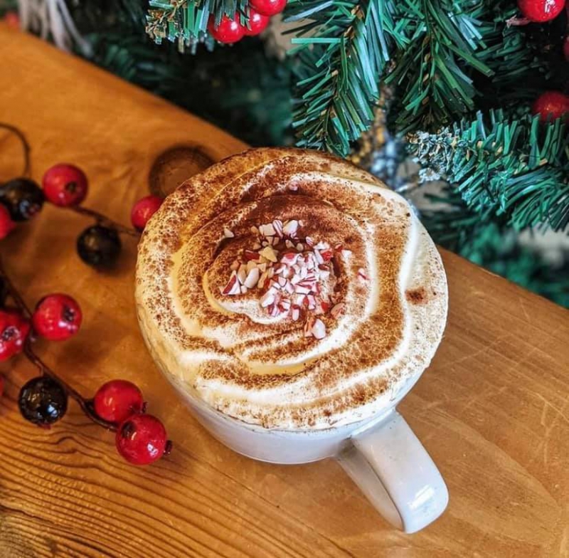 A white mug is topped with whipped cream, dusted with cocoa powder, and topped with crushed peppermints. It sits on a wooden table decorated with red berries on branches. Pine branches are next to the table. Photo from Everyday Kitchen Facebook page. 