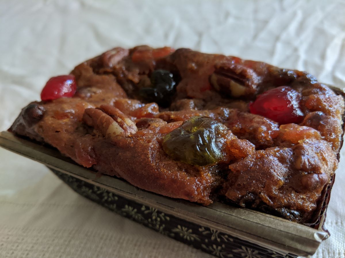 A deep brown loaf cake with whole red and green cherries and pecans. The cake is in a brown paper pan with light brown flowers. The cake is sitting on a beige, wrinkled tablecloth. Photo by Tias Paul.