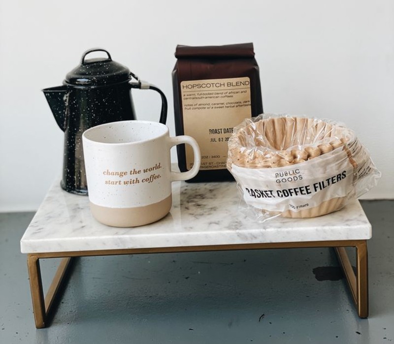 A ceramic tray holds a dark brown bag of coffee, a black coffee pot, a white and light brown coffee mug , and a stack of coffee filters. Photo from Hopscotch Bakery + Market Facebook page. 