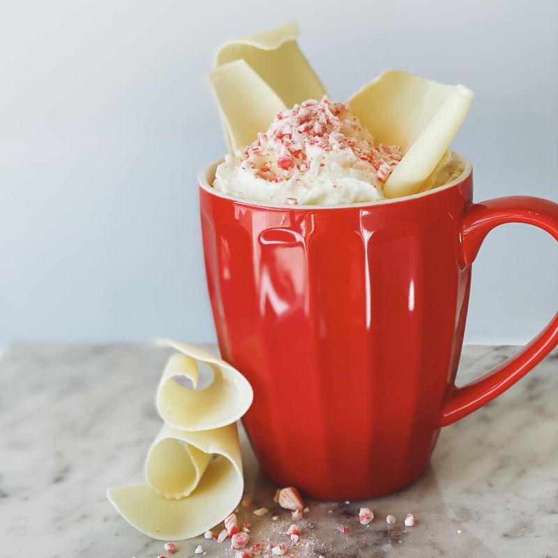 A red mug is topped with whipped cream, crushed peppermint candy, and shavings of white chocolate. It sits on a marble counter, and there are more crushed candies and shavings next to the mug. Photo from Hopscotch Facebook page.