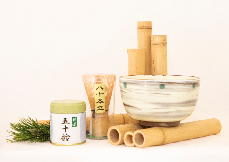 In this photo, the tea beginning set from Japan House is beautifully displayed. There is a jar of matcha, a matcha whisk inside of a plastic container, and a bowl. There are decorative bamboo shoots holding the bowl. Photo provided by Japan House.