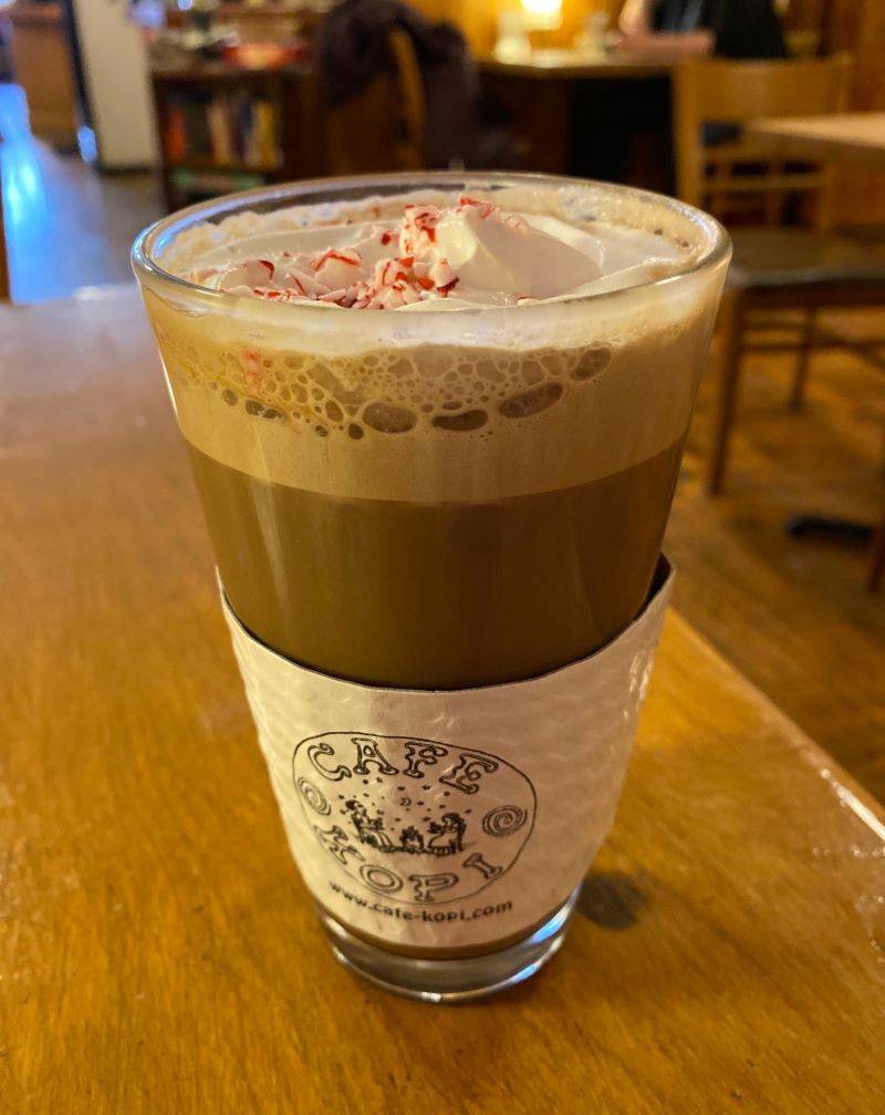 A chocolatey drink is in a tall glass, topped with whipped cream and crushed candy cane pieces. It has a paper holder that says Cafe Kopi, and sits on a wooden table. Photo by Julie McClure.