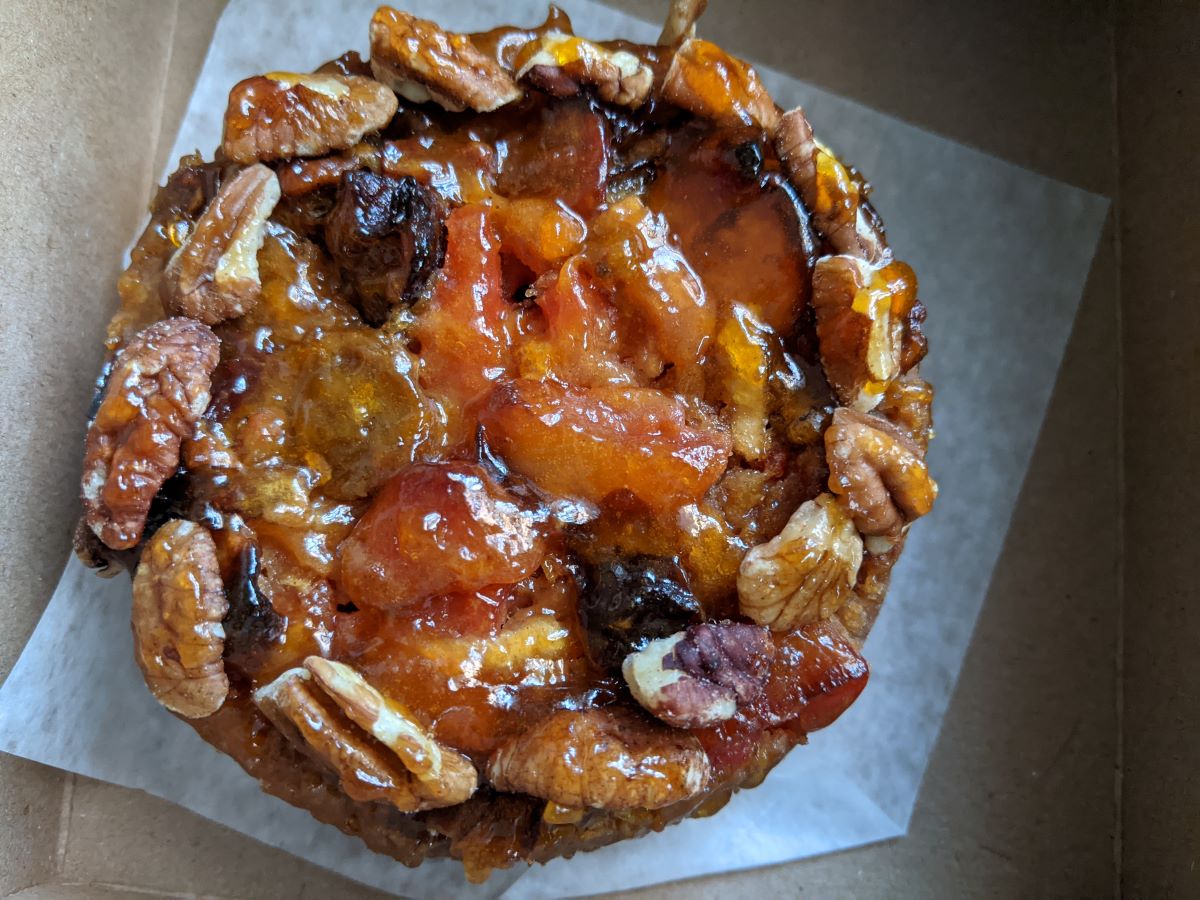 A round cake wreathed in pecans with fruits and shiny fruit preserves in the center.  The cake is sitting on white parchment paper inside a brown cardboard box.  Photo by Tias Paul.