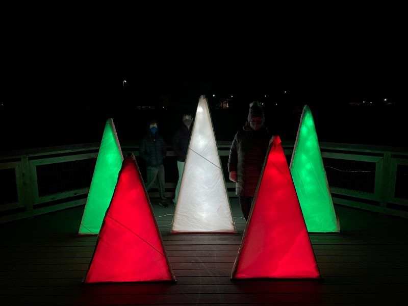 Red, green, and white lighted triangles, representing trees, are sitting on a wooden platform. Photo by Andrew McClure.