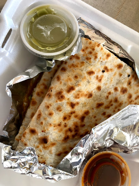A pork quesadilla has light brown grill marks and sits inside a tin foil wrapper. The quesadilla and tin foil are inside a white styrofoam container with two mini salsa containers. Photo by Remington Rock.