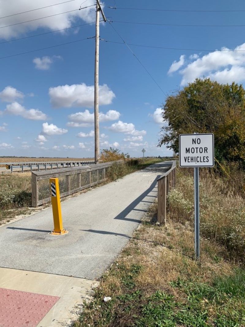 A paved path alongside a road. It's lined by wood fencing, and there is a sign that says No Motorized Vehicles. Photo by Jeff Nelson. 