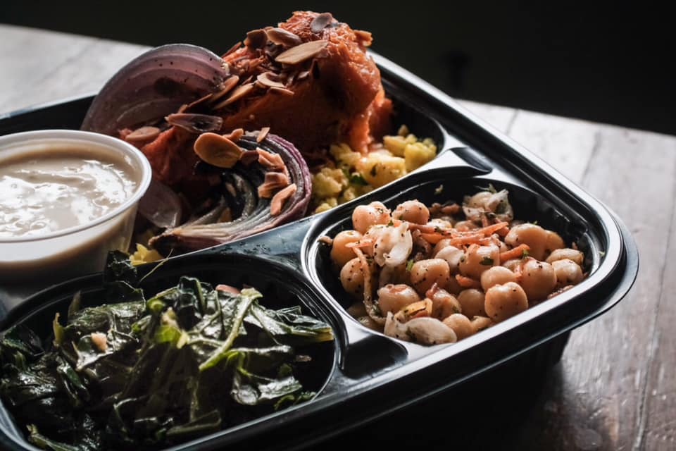 A black divided container has a small cup of brown sauce, a side salad, sauteed greens, and a chickpea salad. Photo from Red Herring's Facebook page.