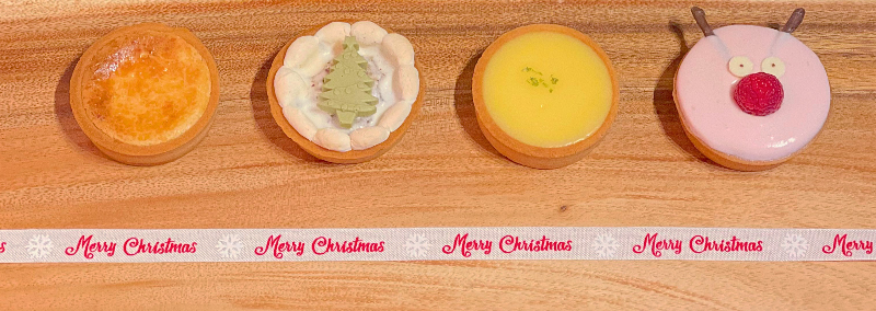 Four tarts sit on a wooden platter from Tasty Tart in Champaign. There is a cheese tart, a s'more tart with a green christmas tree on top of white frosting, a lemon tart, and a raspberry tart shaped like a reindeer. Photo by Tasty Tart.