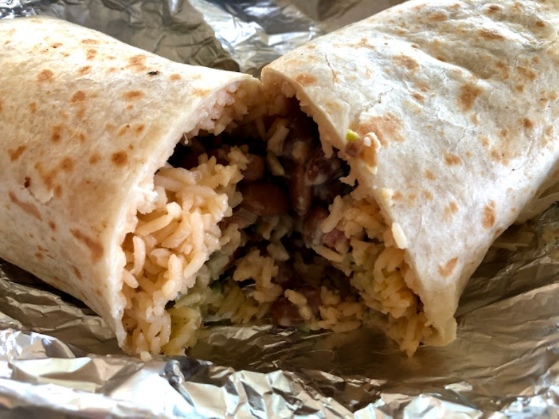 A sliced open burrito reveals rice, beans, and meat inside, sitting on a tin foil paper. Photo by Remington Rock.