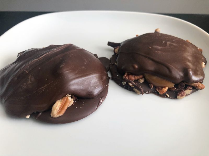 On a white plate, two chocolate turtles from Sugga Shaii's Sweets are next to each other. There is a portion of the pecans under the chocolate poking out. Photo by Alyssa Buckley.