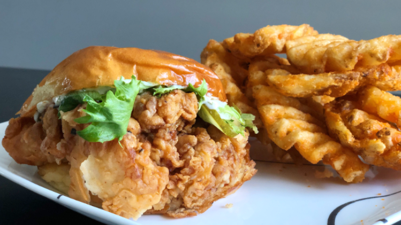Watson's fried chicken sandwich sits on a white plate next to waffle fries. The chicken is battered and coming out of the sandwich with lettuce on top and also coming out of the sandwich. Photo by Alyssa Buckley.