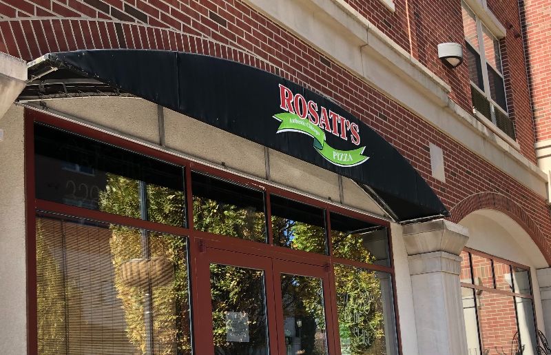 A cropped exterior photo shows the top of the now permanently closed Rosatiâ€™s Pizza in Urbana. The building is red bricked, and the awning reads Rosatiâ€™s in red block letters. The glass windows reflect the green leaves of the trees on Nevada Street. Photo by Alyssa Buckley.