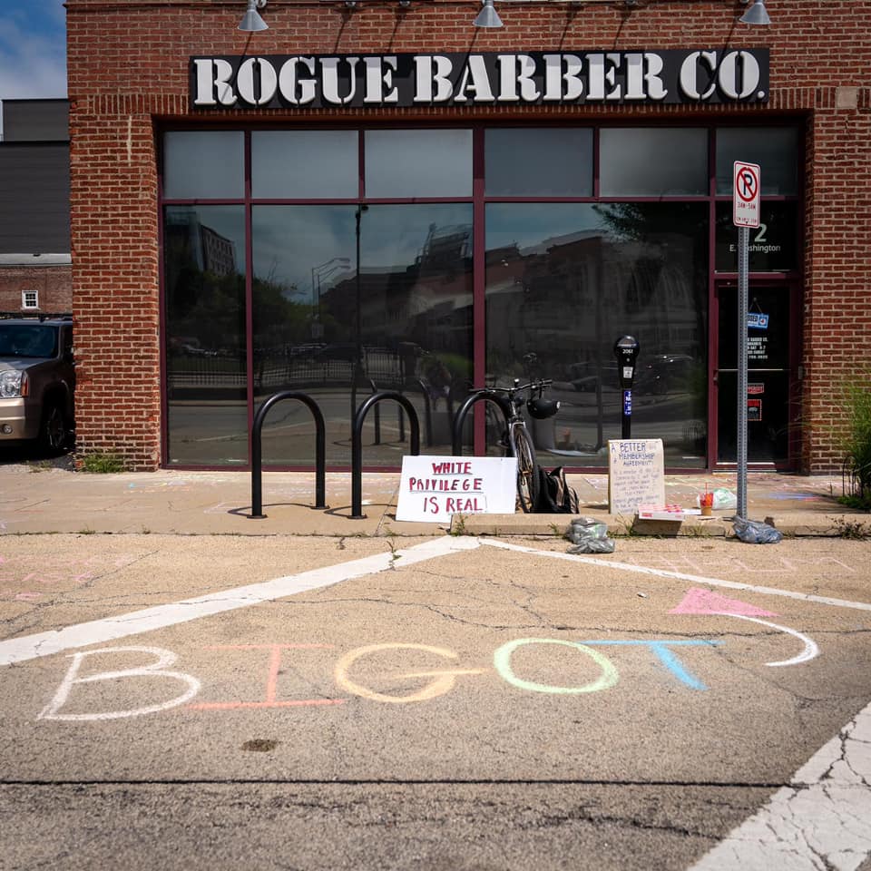 View of the front of Rogue Barber Co, a brick building, from the street, where there are chalk letterings of BIGOT pointing to the front of the business.